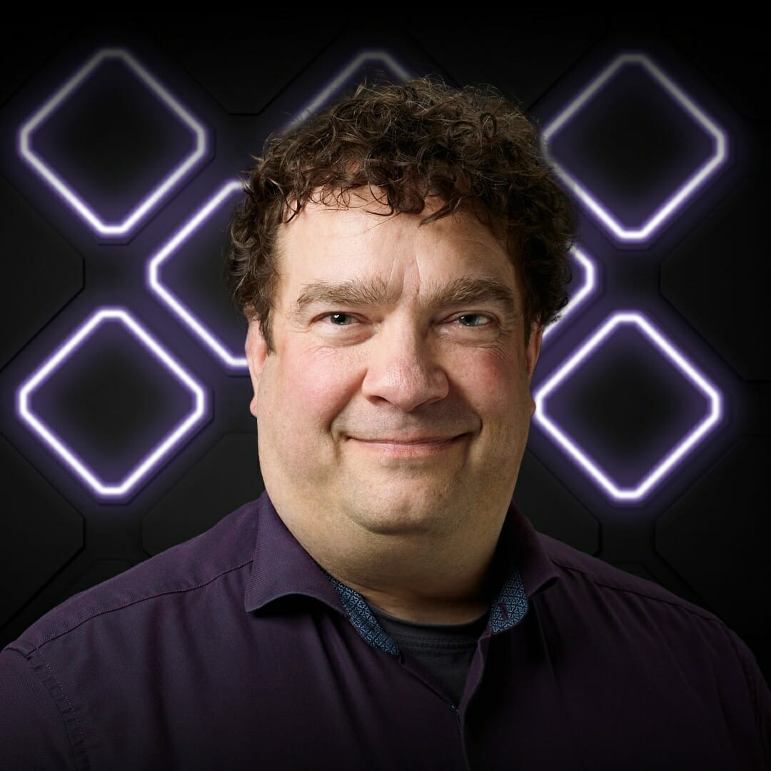 A man with curly dark hair and a slight smile stands in front of a black background adorned with illuminated geometric shapes, capturing the vibrant energy of Spryker EXCITE 2022. He is wearing a dark purple shirt.