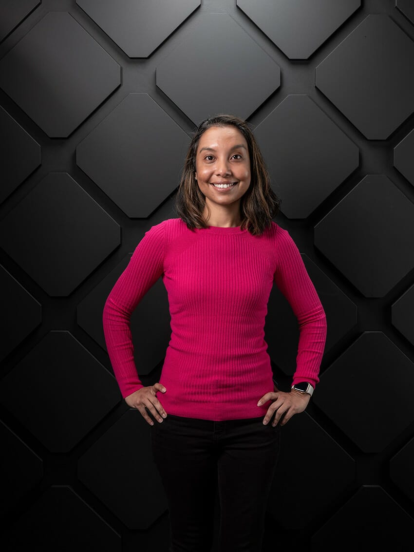 A woman in a bright pink sweater stands with her hands on her hips, smiling, in front of a geometric black wall at Seamless Middle East 2023.