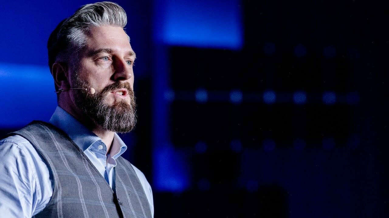A man with a beard and gray hair, wearing a vest and white shirt, appears to be speaking into a microphone headset about the latest Spryker advancements. The background is dark with blue lighting.