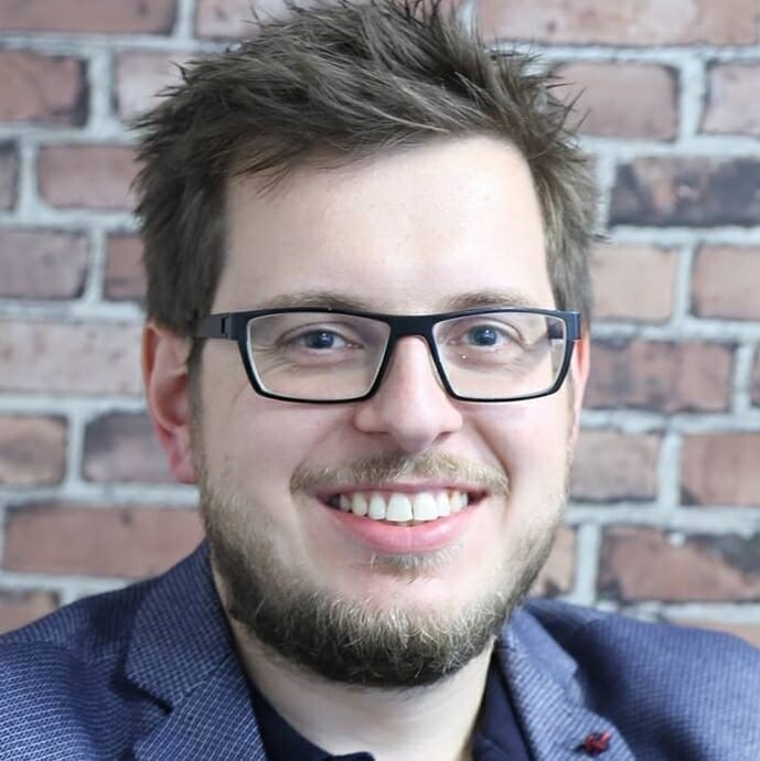 A man with glasses and a beard is smiling. He is wearing a blue jacket and standing in front of a brick wall background, ready to discuss the nuances of SEO.