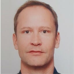 A headshot of a man with short hair and a neutral expression, wearing a dark top, against a plain background, ideal for featuring in customer advisory board presentations.