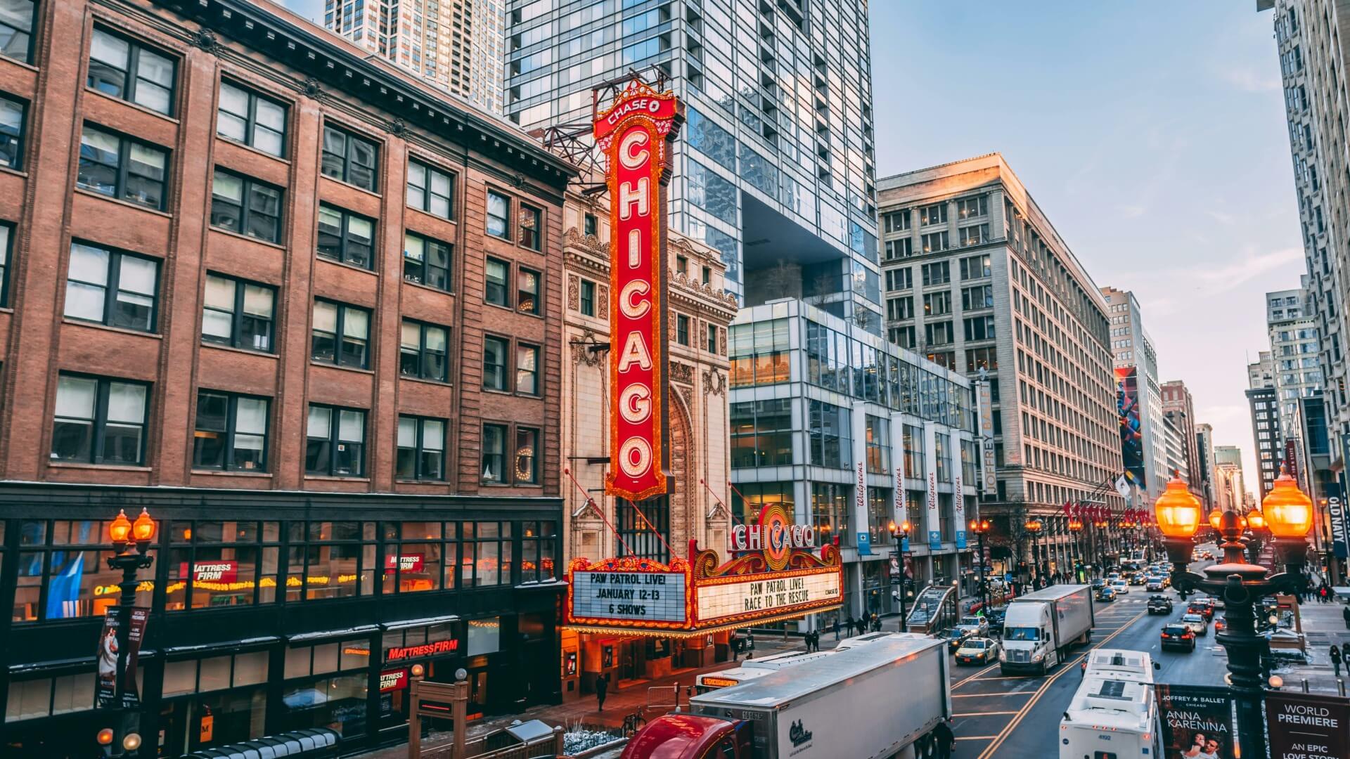 A bustling street in downtown Chicago features the iconic Chicago Theatre marquee sign, lit up in red and white. Surrounding buildings rise high with a mix of historic and modern architecture. The street is busy with cars, trucks, and pedestrians during daylight.