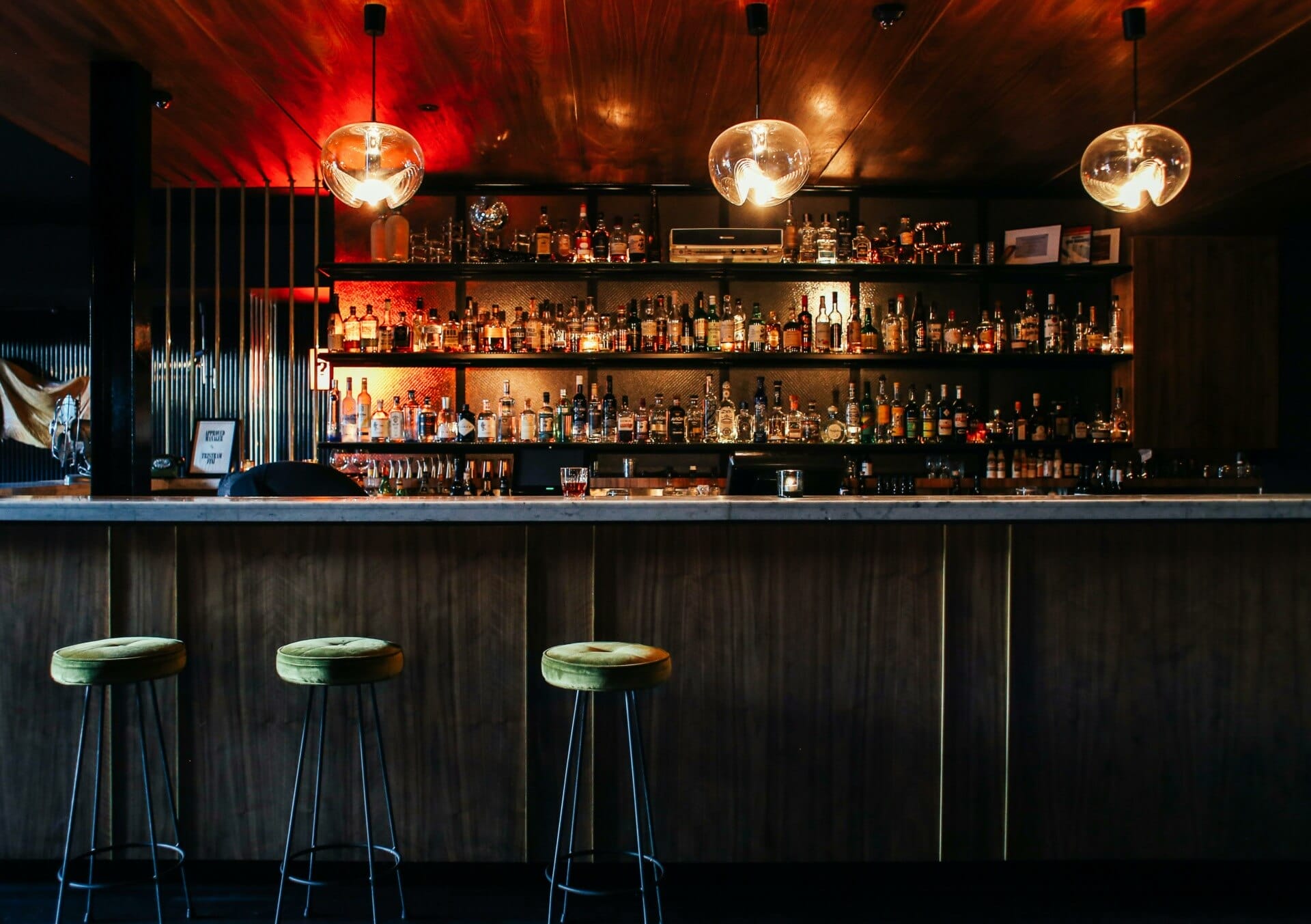 A dimly lit bar with a wooden countertop and shelves lined with various bottles of alcohol. Three yellow cushioned barstools are positioned in front. Hanging light fixtures with exposed bulbs add a warm glow to the scene. The background includes dark wooden and metallic accents.