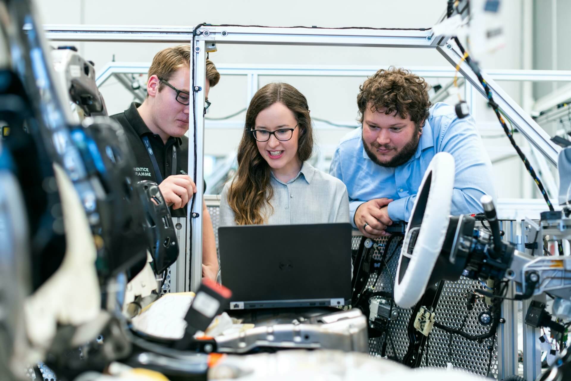 Three individuals in a tech lab work together on a laptop, surrounded by industrial equipment and machinery. The setting appears to be a workshop or a research facility focused on engineering or automotive projects.