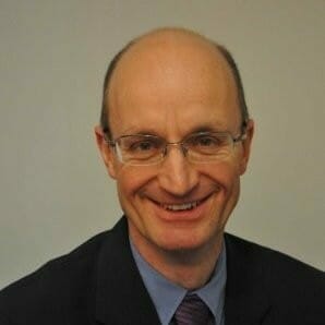 A bald man with glasses and a slight smile, wearing a dark suit jacket, a blue shirt, and a purple tie, is pictured against a plain background. He looks like he's ready to join the customer advisory board.