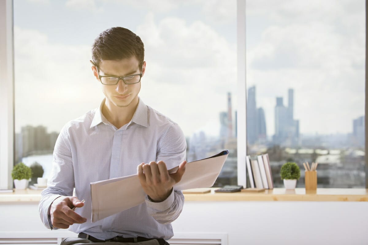 A man wearing glasses and a white shirt sits by a window with a cityscape view, holding and reading a Spryker Raizor folder.
