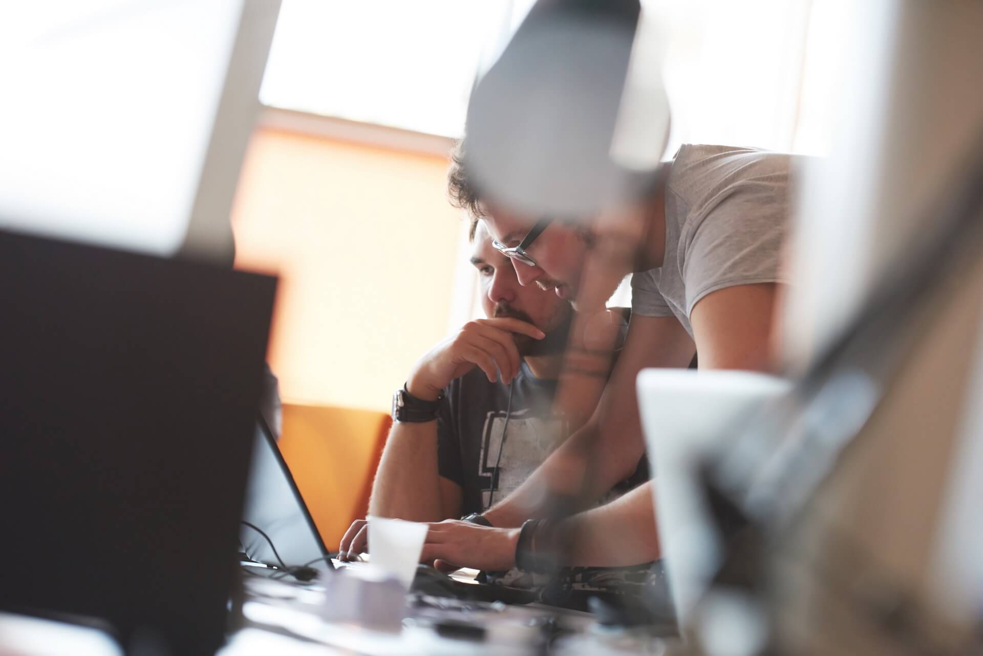Two individuals, partially obscured by blurred foreground objects, working closely together on a laptop in a well-lit room. One is seated and focused on the screen, while the other leans over, pointing at the laptop with apparent concentration.