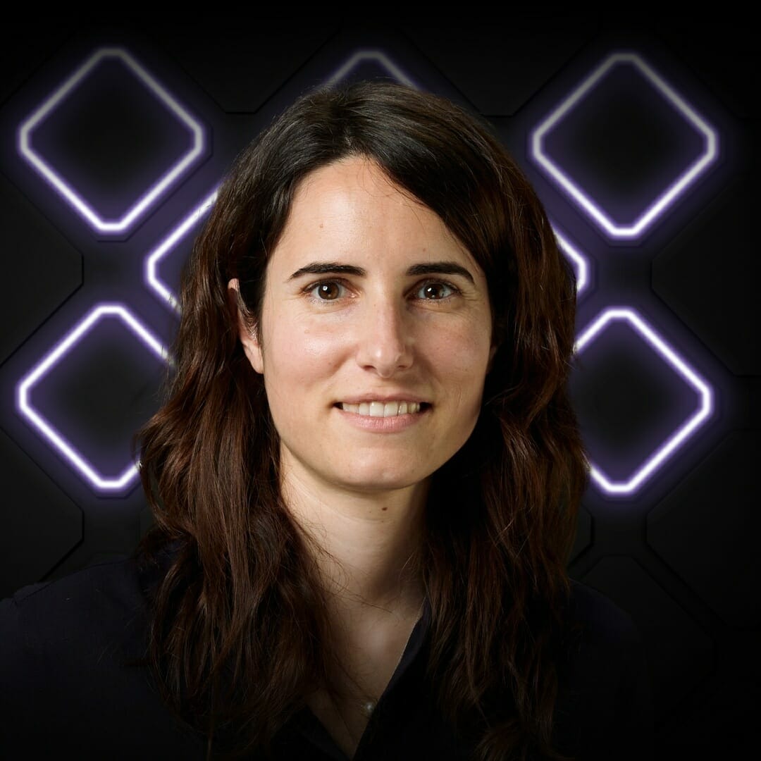 A person with long, dark hair stands in front of a dark background with illuminated geometric shapes at Spryker EXCITE 2022. They are smiling and wearing a dark top.