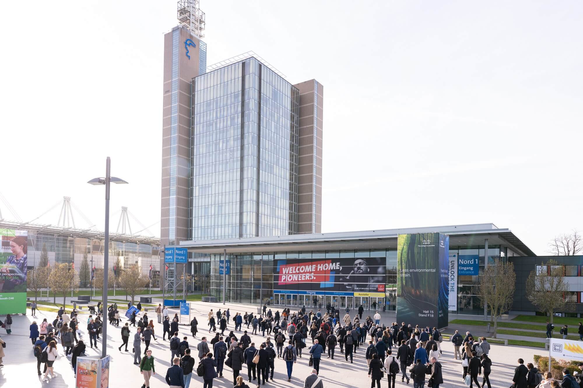 A large group of people walk towards the entrance of a modern building with a tall glass facade. Signs on the building read "WELCOME HOME, PIONEERS." The sky is clear, and there are banners and signs around the area.