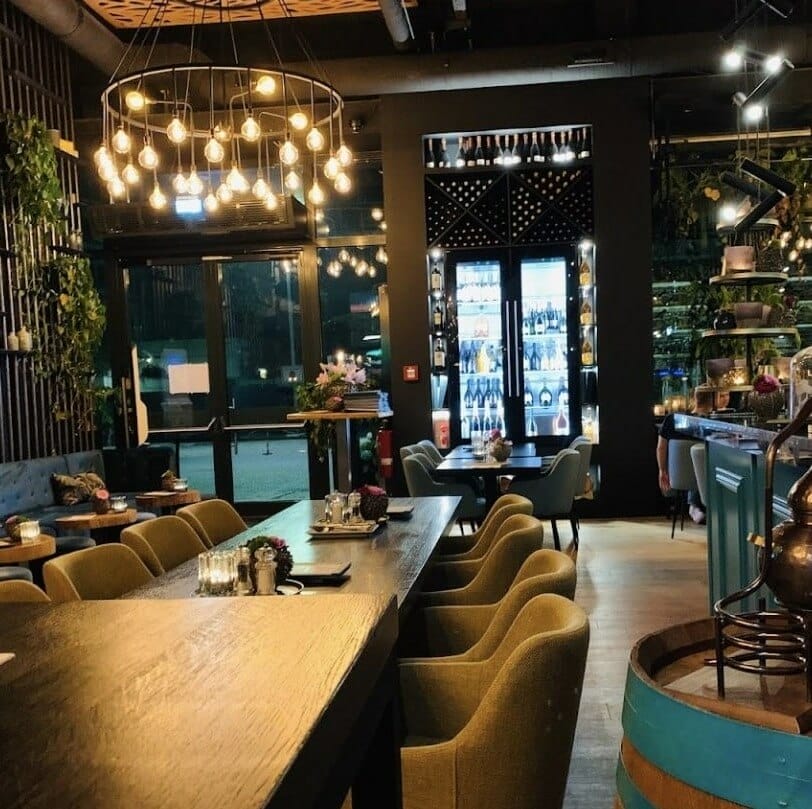 A trendy restaurant with a modern interior design featuring hanging pendant lights, cozy green chairs around dark wooden tables, and a bar area with a variety of bottles. Plants and decorative elements enhance the ambiance, creating a welcoming atmosphere.