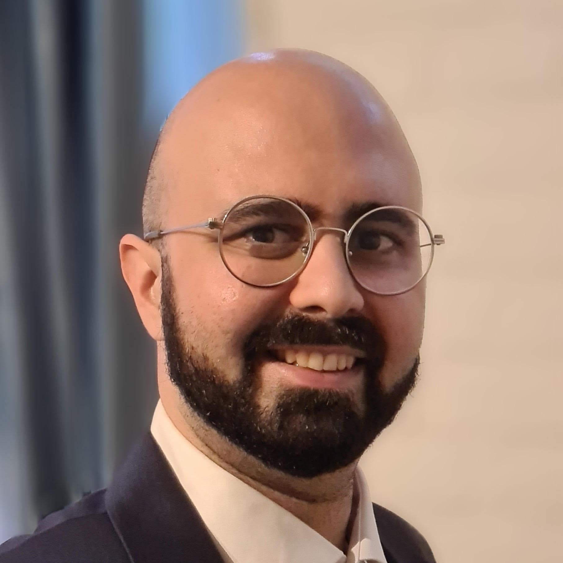 A man with a bald head, glasses, and a beard, wearing a suit, smiles at the camera against a blurred background at Spryker EXCITE 2022.