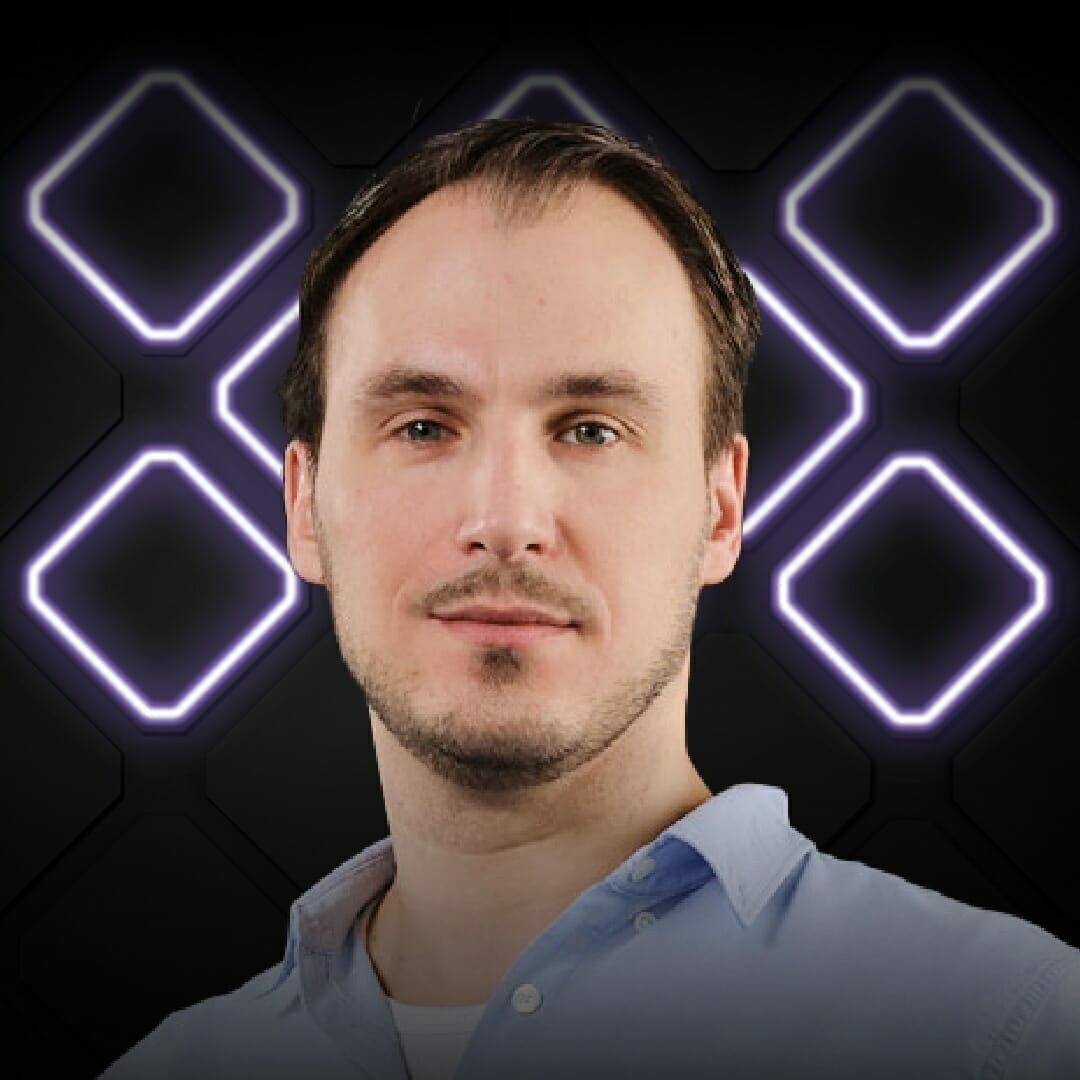 A man with short hair and a trimmed beard, wearing a light blue shirt, stands in front of a dark background illuminated with geometric shapes, capturing the energetic vibe of EXCITE 2022 Nashville.