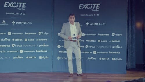 A man in a grey suit speaks on stage at the Excite event in Nashville. The backdrop features various sponsor logos, including Luminos.Labs, PairEye, Vertex, and others, reflecting their strong relationship with Spryker customers.