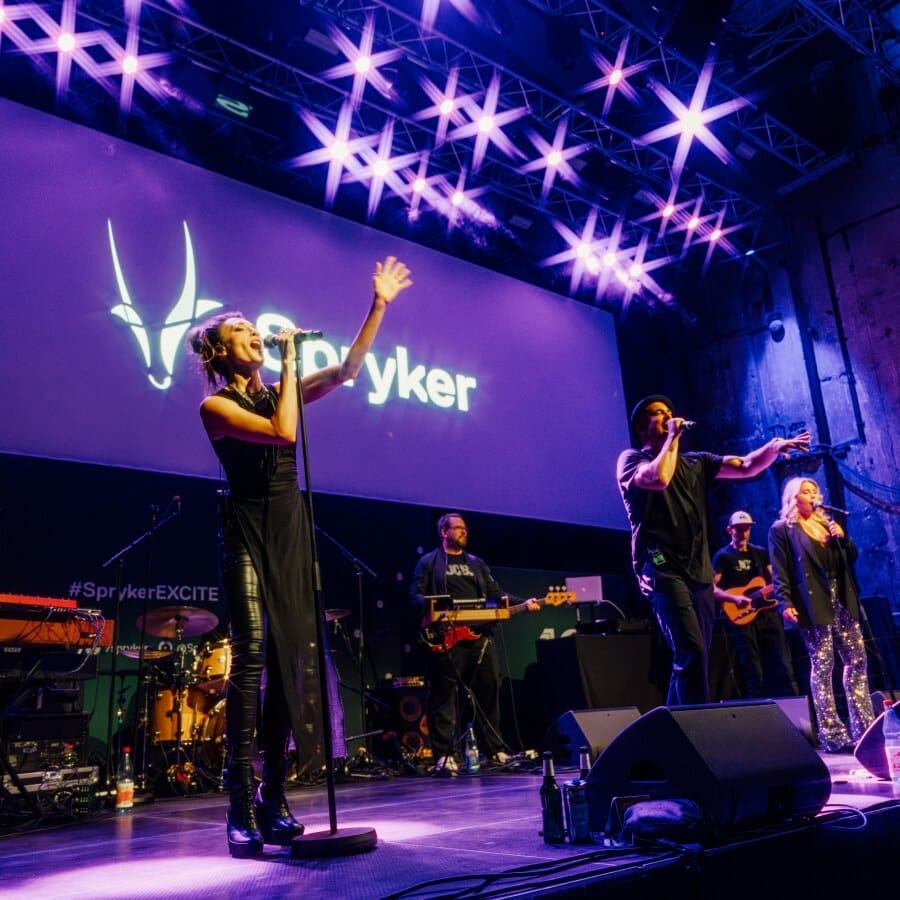 A band performs on stage under purple lighting with a large "Spryker" logo displayed on the screen behind them at Spryker EXCITE 2025.