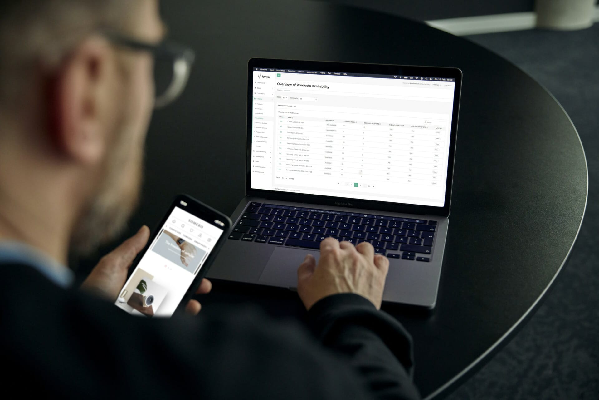 A person using a smartphone and a laptop simultaneously at a round table. The laptop screen displays a Trevipay spreadsheet with product availability data.