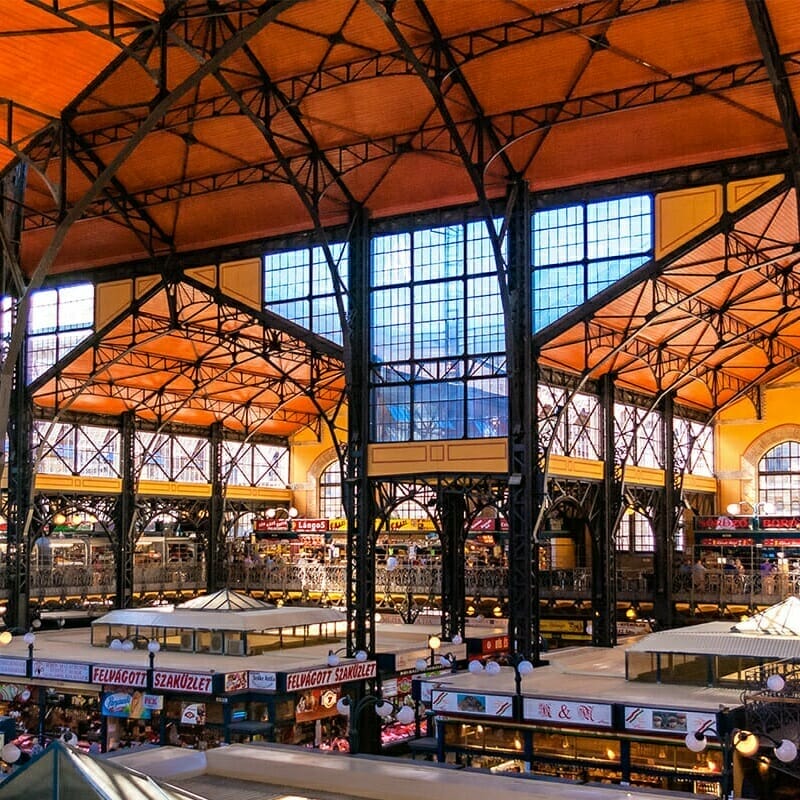 Indoor market hall with high ceilings and iron framework, featuring various stalls and kiosks selling food and goods. Large windows allow natural light to illuminate the greenfield-inspired space.