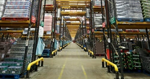 A wide aisle in a large warehouse is shown, lined with tall shelves stacked with various industrial goods and products. Yellow safety barriers and category labels are visible on the shelves.