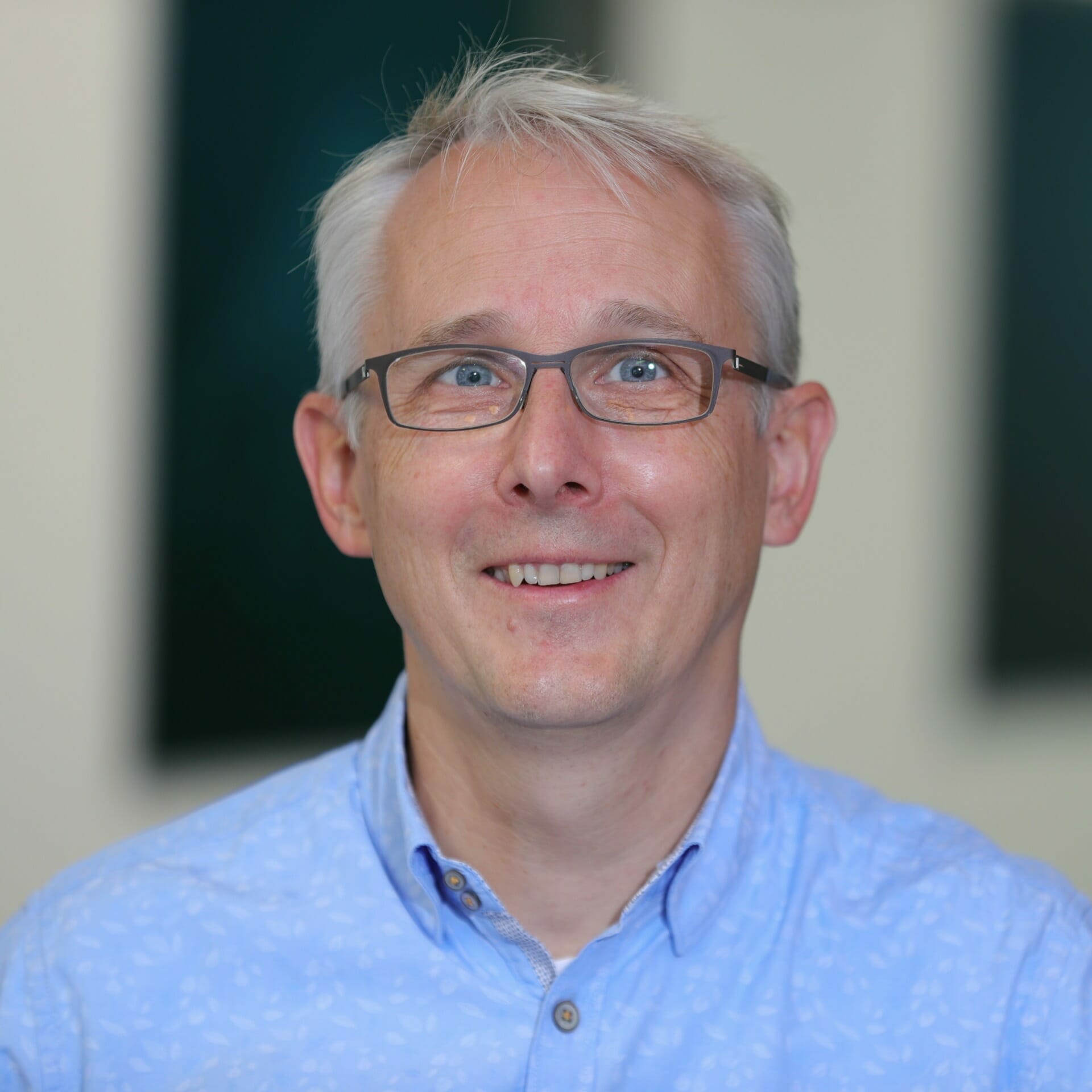 A man with short gray hair and glasses, wearing a light blue button-up shirt, smiles in a well-lit indoor setting during Spryker EXCITE 2022.