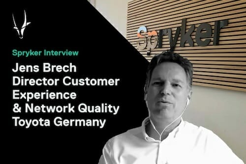A man, identified as Jens Brech, Director of Customer Experience & Network Quality at Toyota Germany, is being interviewed by Spryker. He is wearing a white shirt and earphones, with a Spryker logo visible behind him. The discussion centers around enhancing the journey for Spryker customers.