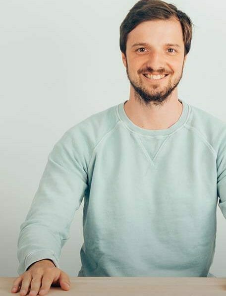 A man with short hair and a beard, wearing a light blue sweatshirt, is sitting at a table, smiling at the camera as if he's ready to discuss the step-by-step replatforming process.