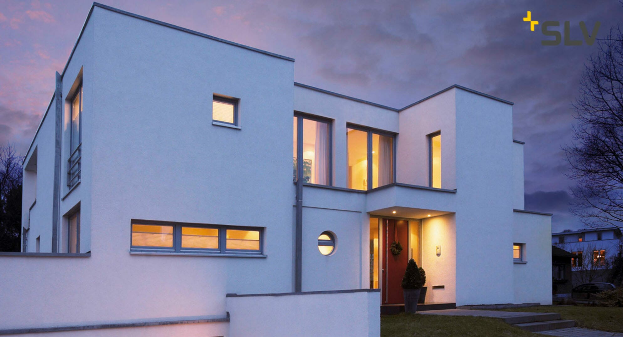 Modern, white, two-story house with large windows, flat roof, and minimalistic design, seen during twilight. The house is lit from within, creating a warm glow against the fading daylight – resembling the sleek headquarters of a digital company.