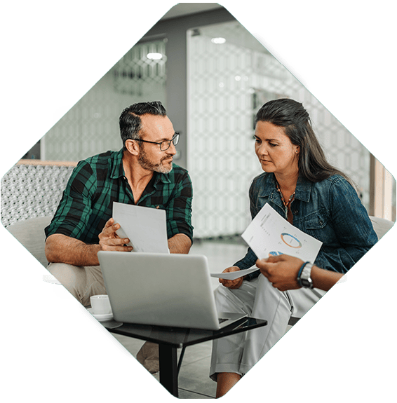 Two people are sitting at a table with a laptop and papers, engaged in discussion about composable commerce. A third person is handing them more documents.