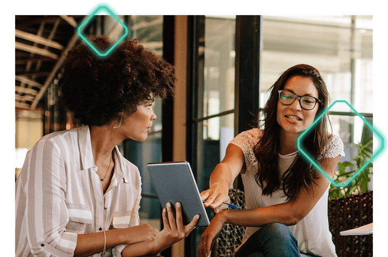 Two women are having a discussion about B2B order management while one points to a tablet the other is holding. They are sitting in a modern office space.