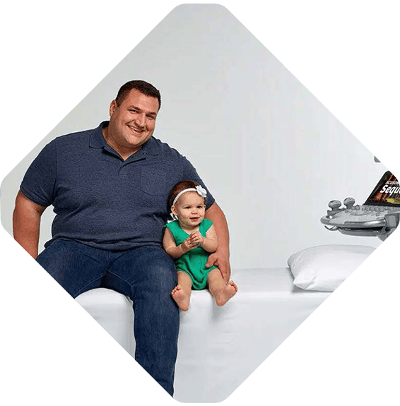 A man sits on a bench next to a smiling baby girl in a green dress and headband, while Siemens Healthineers medical equipment is visible near the bench.