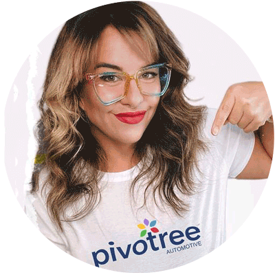 A person with wavy brown hair and colorful glasses smiles and points to their white shirt, which has the word "Pivotree" and "Automotive" printed on it along with a colorful logo.